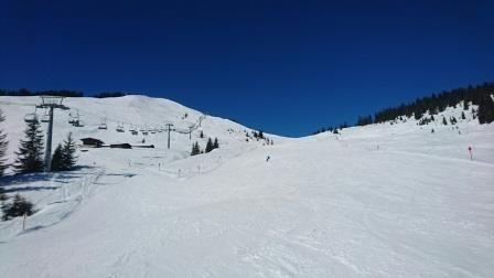 A snow-covered slope under a clear blue sky. In the background, ski lifts and trees are visible.