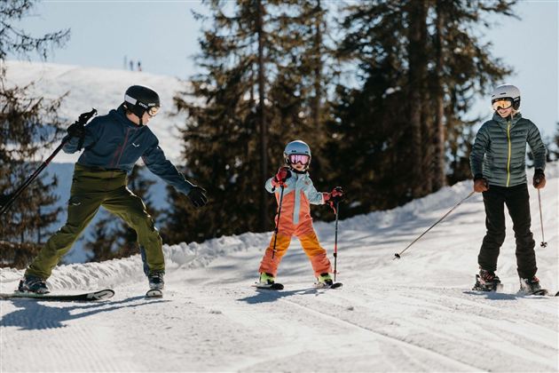 A family is skiing in a sunny winter landscape. The child is learning to ski while the adults watch and help.