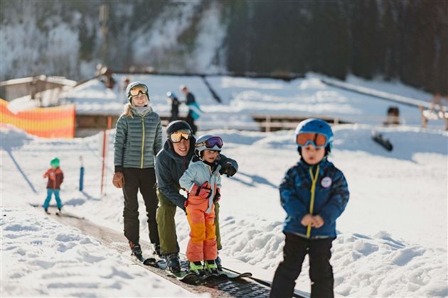 A group of children and adults is standing in a snowy area. They are wearing ski equipment and preparing to go down the slope.