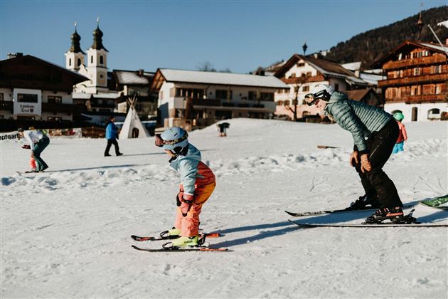A child is learning to ski on a snow-covered slope. In the background, charming alpine huts and churches can be seen.