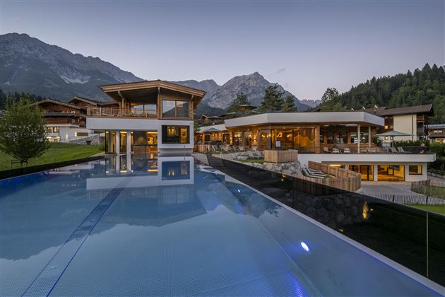 A modern building with large windows and a beautiful infinity pool. In the background, majestic mountains and trees are visible.