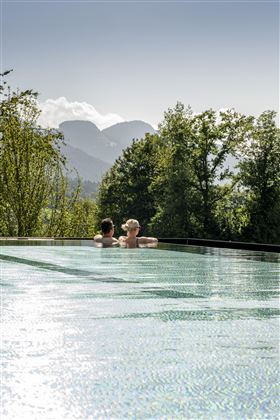 A couple relaxes in an infinity pool overlooking the mountains. The surroundings are green and the sun is shining.