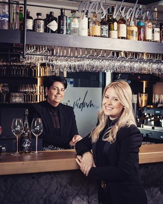 Zwei Frauen stehen hinter einer Bar, umgeben von einer Auswahl an Spirituosen. Beide tragen schwarze Kleidung und lächeln in die Kamera.