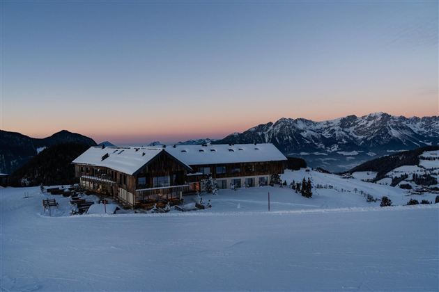 A rustic mountain cabin in the snow in front of majestic mountains. The sky displays soft colors at sunset.