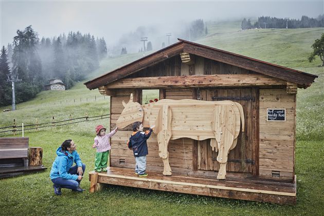 Ein Holzhaus mit einer Holzfigur einer Kuh steht auf einer Wiese. Zwei Kinder und eine Frau stehen davor und erkunden die Figur.