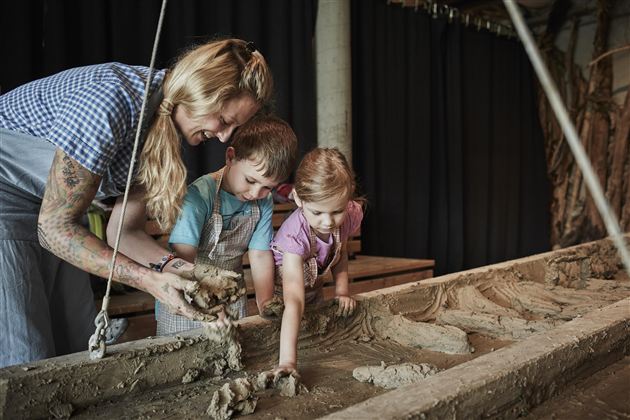 Eine Frau zeigt zwei Kinder, wie man mit Lehm arbeitet. Sie formen gemeinsam etwas in einer Werkstatt.