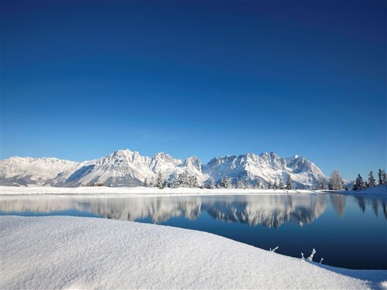 Eine winterliche Landschaft mit schneebedeckten Bergen und einem klaren blauen Himmel. Der ruhige See spiegelt die beeindruckenden Bergen wider.