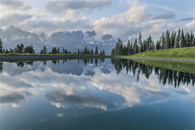 A calm lake reflects the clouds and mountains. Surrounded by trees, the landscape offers a peaceful atmosphere.