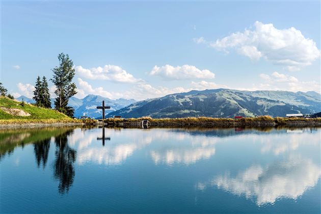 Ein ruhiger See mit klar reflektierten Bergen und einem Kreuz am Ufer. Der Himmel ist blau mit weißen Wolken.