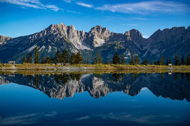 Eine beeindruckende Berglandschaft mit klaren Gipfeln, die sich im ruhigen Wasser spiegelt. Der Himmel ist blau und wolkenlos.