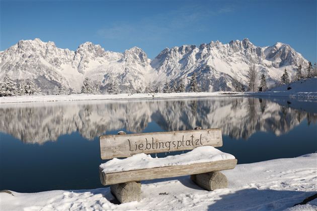 Eine verschneite Landschaft mit hohen Bergen im Hintergrund und einem ruhigen, spiegelnden See. Eine Holzbank mit der Aufschrift "Lieblingsplatz" steht am Ufer.