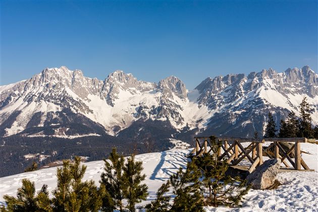 Eine beeindruckende Berglandschaft mit schneebedeckten Gipfeln und klarem blauen Himmel. Im Vordergrund steht ein Holzgeländer, das einen schönen Blick auf die Umgebung bietet.