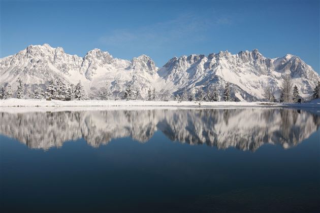 Eine beeindruckende Berglandschaft mit schneebedeckten Gipfeln und einem ruhigen See im Vordergrund. Der klare blaue Himmel spiegelt sich im Wasser.