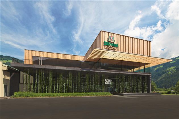 Een modern gebouw met houten bekleding en grote ramen, omringd door groene planten. De lucht is helder en blauw, en het landschap op de achtergrond is bergachtig.