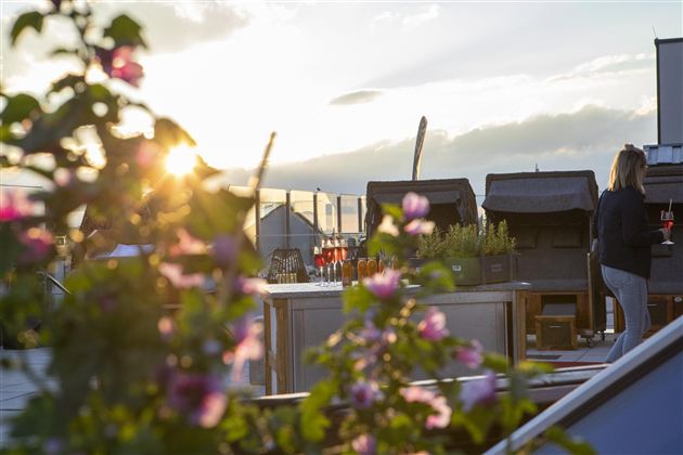 Eine schöne Dachterrasse mit blühenden Pflanzen und einem spektakulären Sonnenuntergang. Im Hintergrund sind gemütliche Liegen zu sehen.