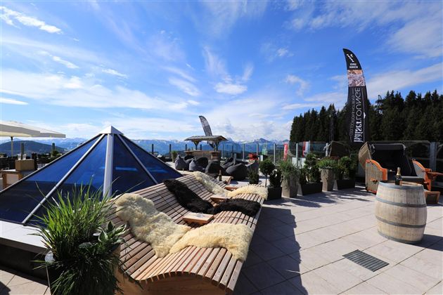 Eine gemütliche Lounge mit Holzliegen und Pflanzen, umgeben von einer atemberaubenden Berglandschaft. Der Himmel ist klar und die Aussicht lädt zum Entspannen ein.
