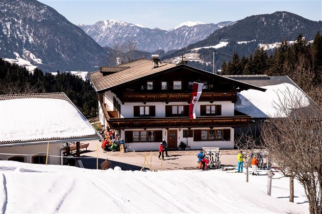 Een traditioneel Alpenhuis ligt in een besneeuwd landschap. Skiërs en gasten zijn op het terrein aan het bewegen, terwijl de bergen op de achtergrond zichtbaar zijn.