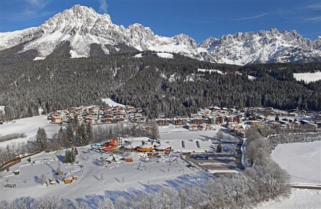 Eine malerische Winterlandschaft mit schneebedeckten Bergen und einem kleinen Dorf. Im Vordergrund sind Freizeitaktivitäten im Schnee zu sehen.