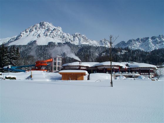 Eine verschneite Winterlandschaft mit hohen Bergen im Hintergrund. Im Vordergrund sind Gebäude und eine Wasserrutsche sichtbar.