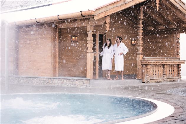 Ein gemütliches Blockhaus im Schnee mit einem entspannenden Whirlpool. Zwei Personen in Bademänteln stehen draußen und genießen die winterliche Atmosphäre.
