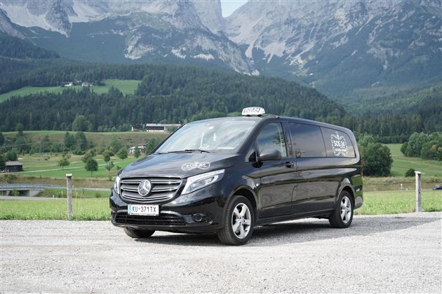 Ein schwarzer Van von Mercedes-Benz steht auf einer gravelartigen Fläche vor einer malerischen Berglandschaft. Im Hintergrund sind grüne Wiesen und hohe Berge sichtbar.