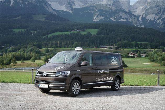 Ein brauner Volkswagen-Taxi-Van steht auf einem Parkplatz mit einer malerischen Berglandschaft im Hintergrund. Die Umgebung ist grün und hügelig unter einem klaren Himmel.