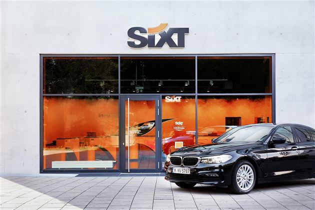 Ein modernes Mietwagenbüro von Sixt mit großen Fenstern. Vor dem Gebäude parkt ein schwarzes Auto.