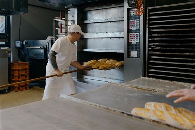 Een bakker haalt vers gebakken brood uit een oven. Op de voorgrond zijn enkele broden en bakgerei te zien.