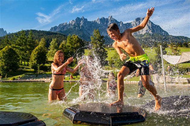 Een groep kinderen speelt in het water en spettert rond. Op de achtergrond zijn bergen en groene bomen te zien.
