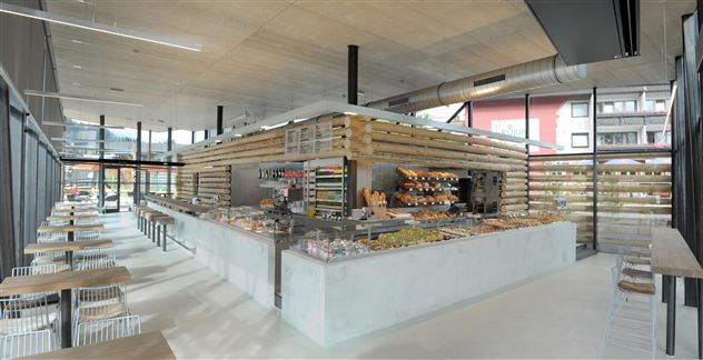 Ein modernes Café mit hellen, offenen Räumen und einer großen Theke, die verschiedene Lebensmittel anbietet. Die Einrichtung besteht aus Holz und bietet eine einladende Atmosphäre.