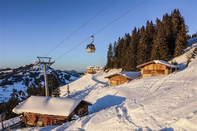 Ein malerisches Winterland mit schneebedeckten Hütten und einer Seilbahn. Hohe Tannenbäume umgeben die Szene, während die Sonne am klaren Himmel scheint.