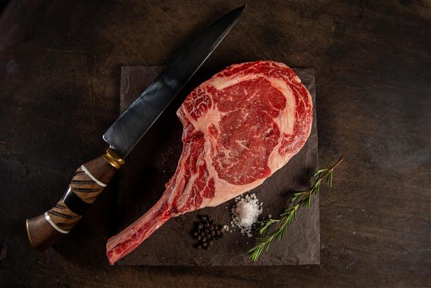 Een rauwe steak met goed zichtbare marmeringen ligt op een zwart bord. Naast de steak bevindt zich een mes en kruiden zoals zout en peper.