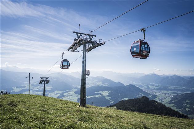 A cable car travels over a green meadow with a view of the mountainous landscape. The sky is clear and blue.