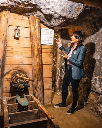 Eine Frau steht in einem Bergwerk und erklärt eine Informationstafel. Um sie herum sind Holzelemente und Bergbaugeräte sichtbar.