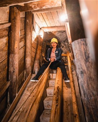 Eine Person sitzt in einem Minenschacht auf einer Holzrutsche und trägt einen gelben Helm. Der Raum ist aus Holz und beleuchtet, was eine spannende Atmosphäre schafft.