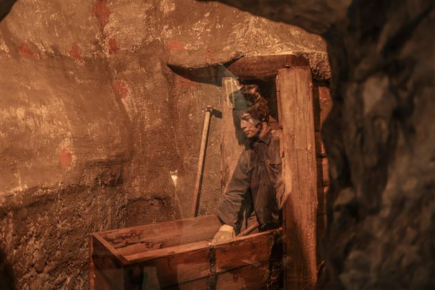 Ein Bergmann arbeitet in einem engen Tunnel. Im Hintergrund sind Werkzeuge und eine Holzkiste zu sehen.