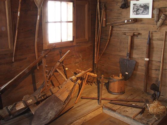 Eine alte Werkstatt mit verschiedenen landwirtschaftlichen Werkzeugen. Die Wände sind aus Holz und ein Fenster lässt Licht herein.
