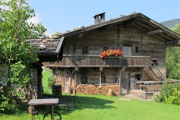 Ein traditionelles Holzhaus mit einem Balkon und roten Blumen. Der Garten ist grün und gepflegt, umgeben von Bergen.