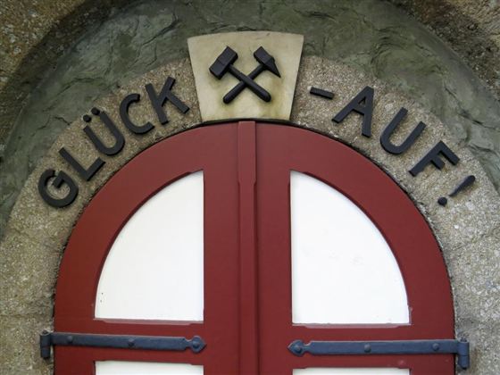 Das Bild zeigt eine rote Tür mit der Aufschrift „GLÜCK -AUF!“ und zwei gekreuzten Hämmern darüber. Die Tür ist Teil eines historischen Gebäudes und vermittelt ein handwerkliches Ambiente.