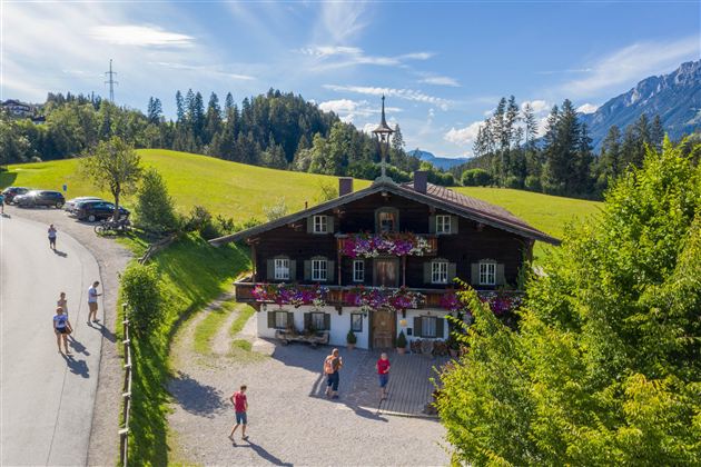 A picturesque alpine house surrounded by green meadows and trees. In front of the house, people and cars can be seen, with mountains in the background.