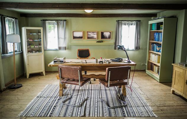 A cozy office with a large wooden desk and two chairs. The walls are painted green, and there are shelves and windows with curtains.
