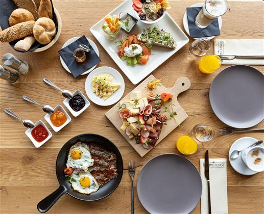 Ein reichhaltiges Frühstücksbuffet mit verschiedenen Speisen, darunter frische Brötchen, Aufschnitt, Rühreier und Obst. Die Tische sind schön gedeckt mit Tellern und Gläsern.