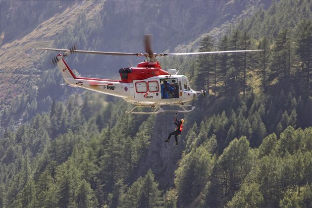 Een helikopter zweeft boven een bos en redt een persoon. De scène toont een heldere lucht en indrukwekkend berglandschap.