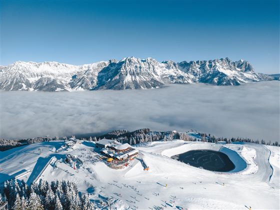 Eine schneebedeckte Berglandschaft mit majestätischen Bergen im Hintergrund. In der Mitte befindet sich eine Hütte, umgeben von Wolken und Skigebieten.