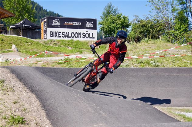 Ein Mountainbiker fährt auf einer kurvigen Strecke. Im Hintergrund ist das Schild "Bike Saloon Söll" zu sehen.