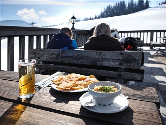 Ein Tisch im Freien mit Blick auf verschneite Berge. Darauf steht ein Glas Bier, ein Teller mit Fladenbrot und eine Schüssel mit warmem Gericht.