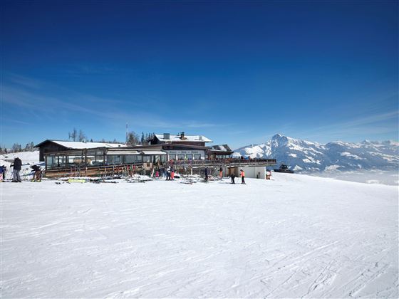 Eine bergige Winterlandschaft mit viel Schnee und klar blauem Himmel. Im Vordergrund steht ein Skihütte, umgeben von Skifahrern.