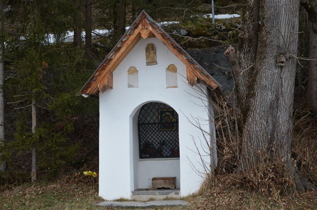 Eine kleine, weiße Kapelle mit einem Holzdach steht im Wald. Umgeben von Bäumen ist sie an einer ruhigen Stelle platziert.