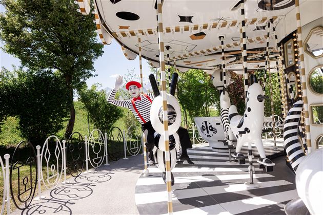 A carousel with a cheerful mime in a striped outfit. The surroundings are green and sunny, featuring a creative design of the attraction.