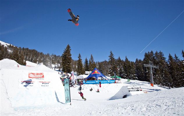 Ein Snowboarder springt in die Luft über einen verschneiten Park. Im Hintergrund sind Bäume und ein Skilift sichtbar.